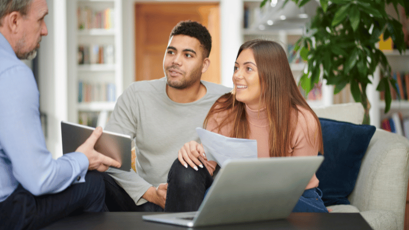 A young couple sitting on a sofa and listening intently to a mortgage advisor who is explaining documents to them.