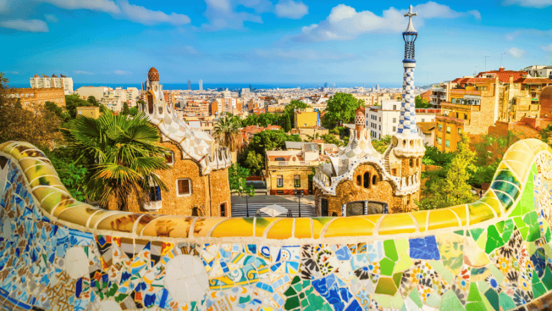 Barcelona skyline from Park G&uuml;ell with Gaud&iacute;&rsquo;s mosaic terrace and historic architecture under a blue Mediterranean sky.