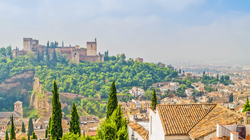 A castle in Granada surrounded by trees with the historic town in the foreground.