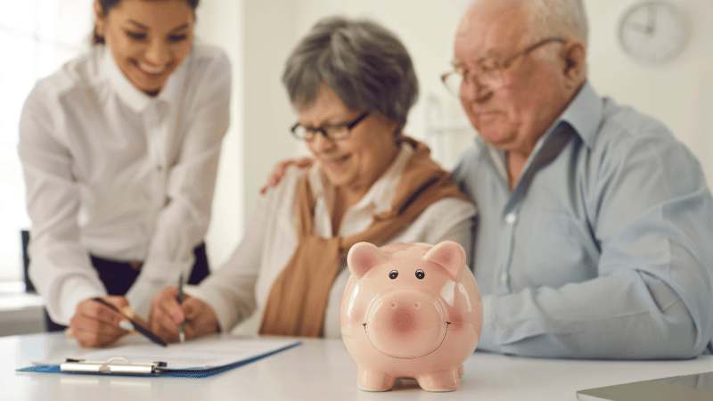 A smiling senior couple signing a pensioner mortgage agreement with a professional female advisor, with a pink piggy bank on the table symbolizing their retirement savings.
