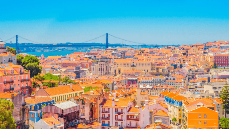A sunny, high-angle view of the orange-roofed cityscape of Lisbon, Portugal, with the 25 de Abril Bridge in the background over the Tagus River.