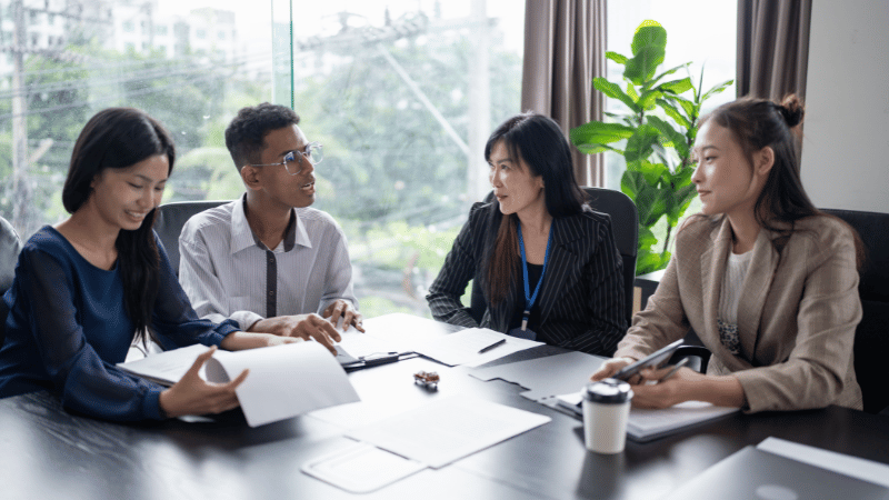 A group of four business professionals collaborating around a boardroom table covered with documents, representing a strategic meeting about securing a business loan for growth.