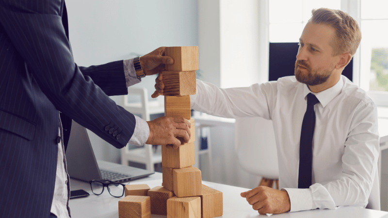 Two business professionals building a wooden block tower representing the steps, timelines, and funding layers of the UK management buyout process.