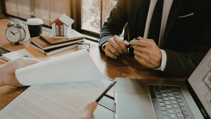 A mortgage broker reviewing documents with a client at a desk with a model house, representing professional advice on the UK property market forecast.